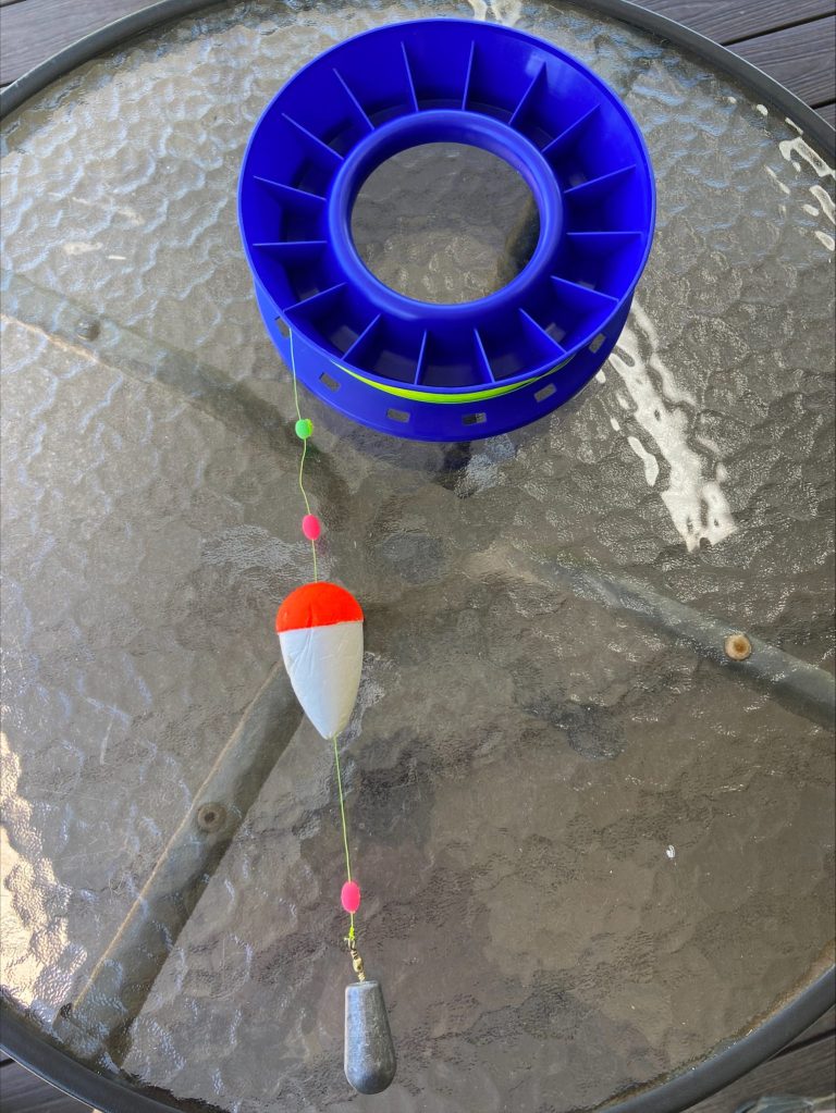 DAMDEEP Farm dam measuring tool, consisting of a blue fishing line spool with a float, beads and a sinker, sitting on a glass table