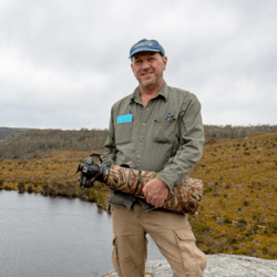 Peter Rowland standing on a rocky ledge with Tasmania's Dove Lake in the background